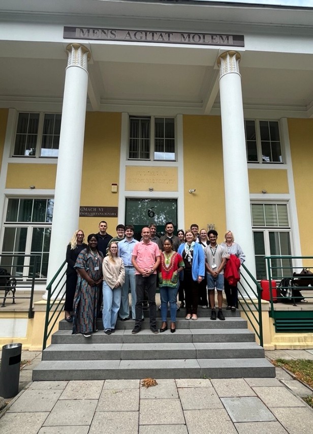 A diverse group of people poses on the steps of a building with columns, featuring a sign above that reads "MENS AGITAT MOLEM."